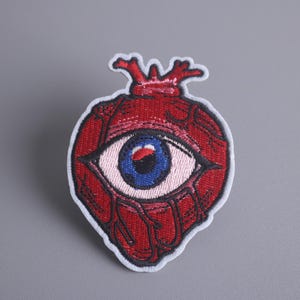 May include: Red embroidered patch of a heart with an eye in the center. The eye is blue with a black pupil and white sclera.
