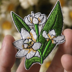 May include: Embroidered floral patch featuring white flowers with yellow centers and green leaves. The patch has a white border and is held in a hand. The background is blurred with other flowers.