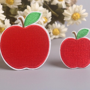 以下が含まれることがあります： 緑の葉と白い縁取りが付いた、赤いリンゴ型の刺繍パッチが2つ。1つのパッチはもう1つよりも大きいです。パッチは、白と黄色の花のぼやけた背景がある灰色の表面にあります。