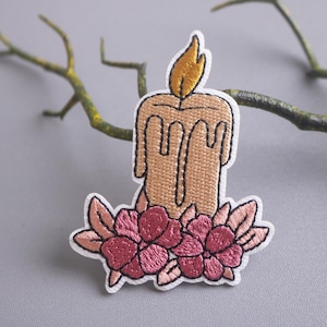 May include: Embroidered iron-on patch featuring a candle with a flame and pink flowers. The candle is a light brown color and the flowers are a light pink color.