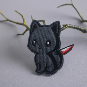 May include: Black embroidered patch of a cartoon cat with a red knife for a tail. The cat has large eyes and a small smile.