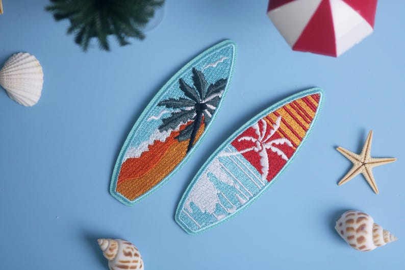 Op de afbeelding: Twee geborduurde surfplankpatches met tropische ontwerpen. E&eacute;n heeft een palmboom over een oranje en geel strand, de andere een rode en gele zonsondergang met een palmboom. De patches zijn lichtblauw omlijnd.