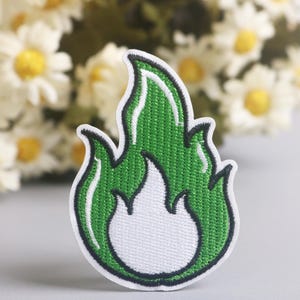 May include: Embroidered patch featuring a green flame design with a white center and black outline. The patch has a textured appearance and is likely for application on clothing or accessories. Daisies are in the background.