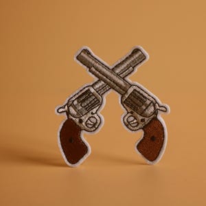 May include: A white embroidered patch of two crossed revolvers with brown grips.