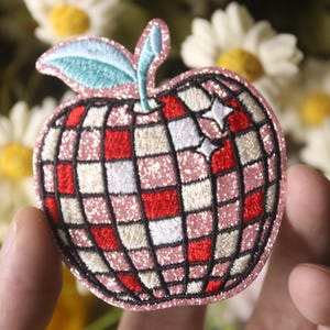May include: A pink and red embroidered iron-on patch shaped like an apple with a disco ball design. The apple has a green leaf and white and red squares.
