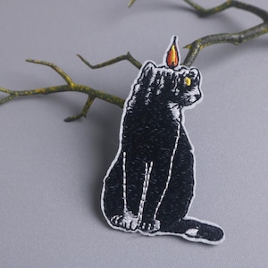 Può includere: Toppa ricamata raffigurante un gatto nero con una fiamma di candela sulla testa. Il gatto è seduto con dettagli di cucitura bianchi. La toppa ha un bordo bianco ed è su uno sfondo grigio con un ramo.