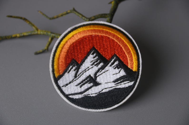 May include: Embroidered patch featuring a mountain range with three peaks in white, set against a sunset gradient of orange and yellow. The patch is circular with a black border, suitable for jackets or bags.