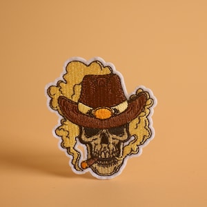 Puede incluir: Parche bordado para planchar con un cráneo que lleva un sombrero de vaquero marrón y fuma un cigarro. El cráneo está rodeado de humo amarillo.