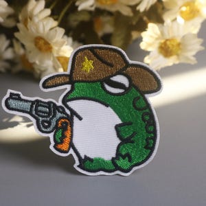 May include: Embroidered patch featuring a green frog wearing a brown cowboy hat with a yellow star and holding a silver revolver. The frog has a white belly and is outlined in black.