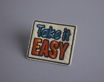 Könnte beinhalten: Ein beigefarbener Stoff-Patch mit schwarzen Nähten an den Rändern. Der Patch hat die Worte "Take it EASY" in blauer und orangefarbener Stickerei.