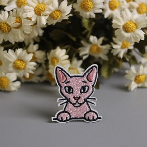 以下が含まれることがあります： キラキラした質感、黒いひげ、黒い輪郭のピンクの猫のパッチ。 パッチは、背景に白と黄色のデイジーのブーケがある灰色の背景に設定されています。