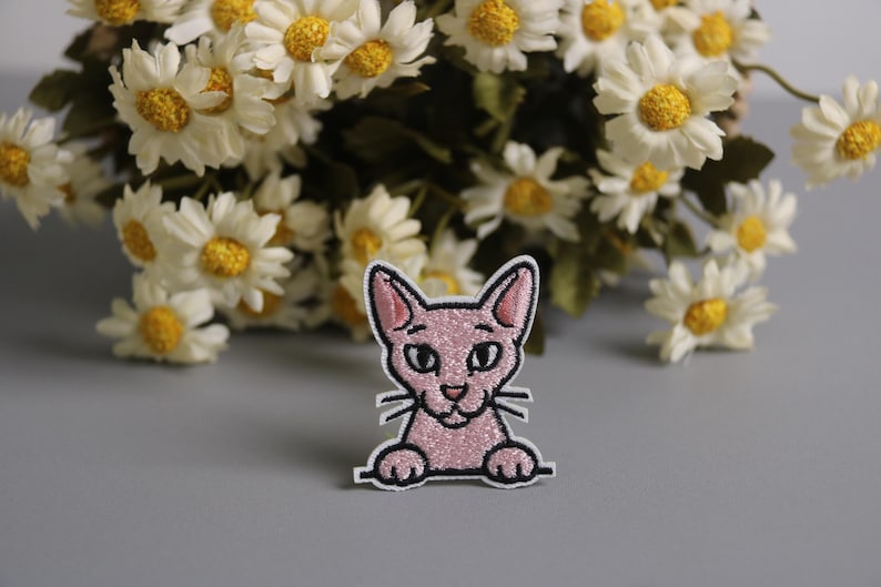 以下が含まれることがあります： キラキラした質感、黒いひげ、黒い輪郭のピンクの猫のパッチ。 パッチは、背景に白と黄色のデイジーのブーケがある灰色の背景に設定されています。