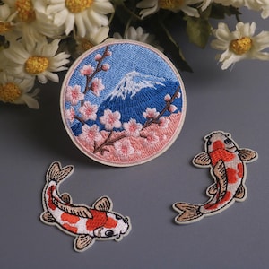 Könnte beinhalten: Gestickte Aufnäher mit japanischen Motiven. Ein runder Aufnäher zeigt den Fuji mit Kirschblüten, und zwei Aufnäher zeigen Koi-Karpfen. Die Aufnäher sind in Blau, Rosa, Weiß und Orange gehalten.