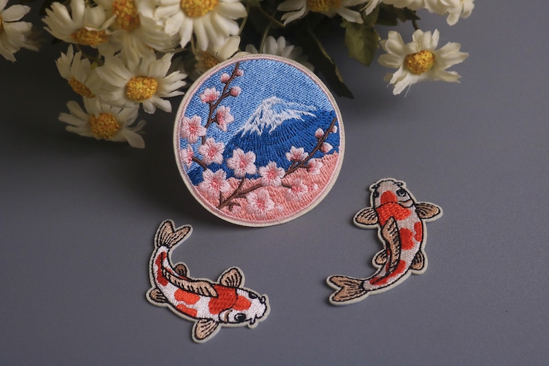 May include: Embroidered patches featuring Japanese motifs. One patch depicts Mount Fuji with cherry blossoms, and two patches show koi fish. The patches are in shades of blue, pink, white, and orange.