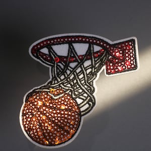 May include: Embroidered patch featuring a basketball going through a hoop. The basketball and backboard are embellished with sequins. The net is black, and the rim and backboard are red. The patch has a white border.