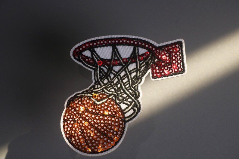 Peut inclure: &Eacute;cusson brod&eacute; repr&eacute;sentant un ballon de basket passant &agrave; travers un panier. Le ballon et le panneau sont orn&eacute;s de paillettes. Le filet est noir, et le bord et le panneau sont rouges. L'&eacute;cusson a une bordure blanche.