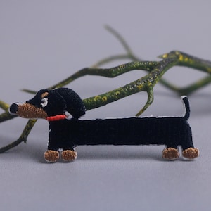以下が含まれることがあります： 黒、茶色、ベージュのダックスフント刺繍パッチ。犬は赤い首輪をしており、緑と黄色の枝のある灰色の背景に配置されています。犬好きのためのキュートなアクセサリーです。