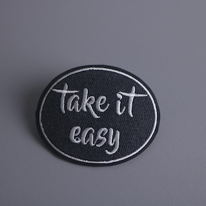 Pode incluir: Um remendo ovalado preto bordado com texto branco que diz "take it easy".