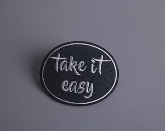 Könnte beinhalten: Ein schwarzer, ovaler gestickter Aufnäher mit weißem Text, der "take it easy" lautet.