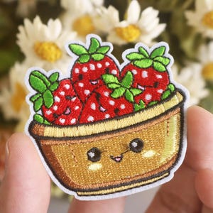 May include: A cute embroidered patch of a bowl of strawberries with a happy face. The bowl is brown and the strawberries are red with green leaves.