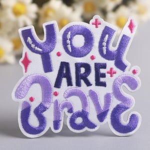 May include: Embroidered patch with the words "You Are Brave" in purple and lavender thread on a white background. The patch features decorative pink accents and a stylized design, suitable for clothing or accessories.