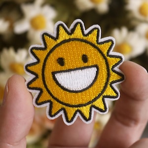 Może przedstawiać: Żółte uśmiechnięte słońce z białym otworem gęby i czarnymi oczami, wyszyte na białym łatce z tkaniny.