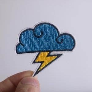 May include: Embroidered patch featuring a blue cloud with a white outline and a yellow lightning bolt. The patch is held by a hand against a white background. This patch is suitable for jackets, bags, or other fabric items.