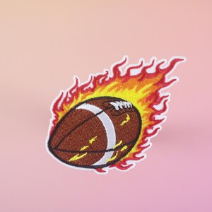 Puede incluir: Un balón de fútbol marrón y blanco en llamas con llamas rojas y amarillas. El balón de fútbol está en el centro de la imagen y las llamas lo rodean.
