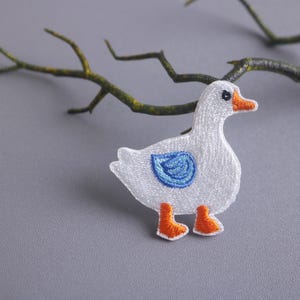 Op de afbeelding: Witte geborduurde ganzen-applicatie met blauw vleugeldetail en oranje poten. De applicatie is op een grijze achtergrond.