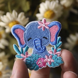 Op de afbeelding: Een geborduurde olifant patch met een blauwe olifant met roze oren en een bloem op zijn kop. De olifant is omringd door groene bladeren en roze bloemen.