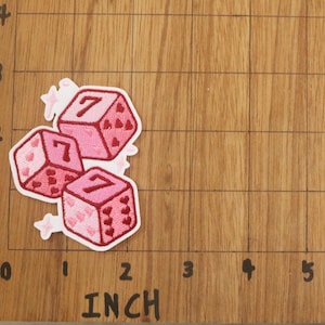 K&ouml;nnte beinhalten: Drei rosa W&uuml;rfel-Patches mit der Zahl 7 auf jeder Seite, verziert mit herzf&ouml;rmigen Augen. Wei&szlig;e Stern-Akzente umgeben die W&uuml;rfel. Die Patches liegen auf einer Holzoberfl&auml;che mit Zollmarkierungen. Die W&uuml;rfel messen etwa 2,5 cm.