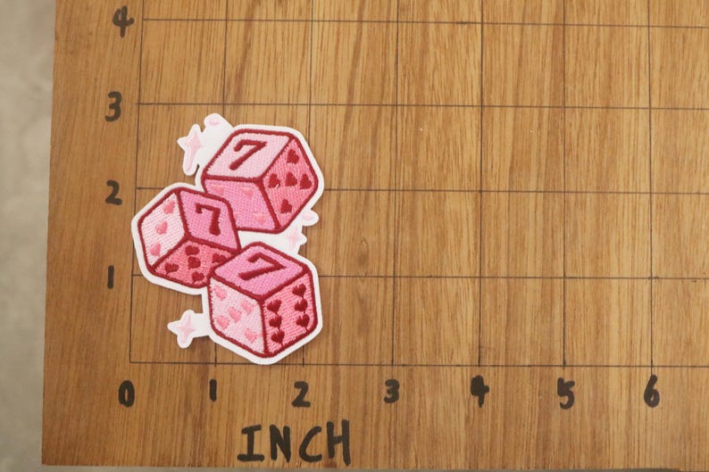 K&ouml;nnte beinhalten: Drei rosa W&uuml;rfel-Patches mit der Zahl 7 auf jeder Seite, verziert mit herzf&ouml;rmigen Augen. Wei&szlig;e Stern-Akzente umgeben die W&uuml;rfel. Die Patches liegen auf einer Holzoberfl&auml;che mit Zollmarkierungen. Die W&uuml;rfel messen etwa 2,5 cm.