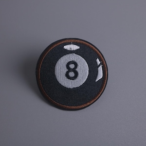 Könnte beinhalten: Ein gestickter Aufnäher einer Billardkugel mit der Nummer 8. Der Aufnäher ist schwarz-weiß mit einem braunen Rand.