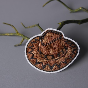 以下が含まれることがあります： 白い縁取りのある茶色のカウボーイハットの刺繍パッチ。帽子は詳細なステッチとクラシックなウエスタンデザインが特徴です。パッチは、緑の枝のある灰色の背景に配置されています。