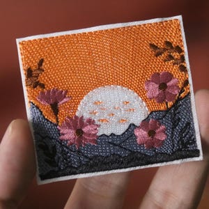 Puede incluir: Parche bordado con una escena de puesta de sol con una luna blanca, un cielo naranja y flores rosas. El parche está sobre un fondo blanco.
