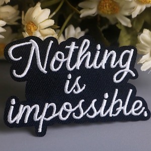 May include: A black embroidered patch with the words "Nothing is impossible" in white cursive script. The patch has a scalloped edge and is set against a backdrop of white and yellow flowers.