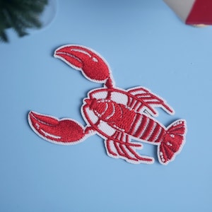 Könnte beinhalten: Gestickter roter Hummer-Aufnäher mit weißen Akzenten, vor hellblauem Hintergrund. Der Aufnäher zeigt eine detaillierte Hummer-Design, ideal, um Kleidung oder Accessoires einen maritimen Touch zu verleihen.