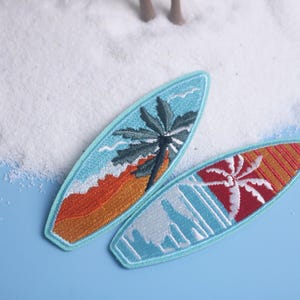 Op de afbeelding: Twee geborduurde surfboard patches. E&eacute;n heeft een palmboom en golven in blauw, oranje en wit. De andere heeft een rood en oranje ontwerp met een palmboom. Beide hebben een lichtblauwe rand.