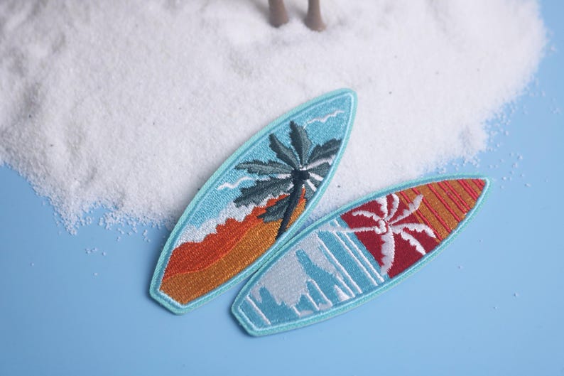 Op de afbeelding: Twee geborduurde surfboard patches. E&eacute;n heeft een palmboom en golven in blauw, oranje en wit. De andere heeft een rood en oranje ontwerp met een palmboom. Beide hebben een lichtblauwe rand.