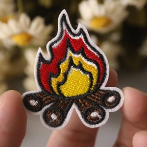 May include: Embroidered iron-on patch depicting a campfire with red and yellow flames. The patch is ideal for adding a touch of rustic charm to clothing, bags, or other fabric items.