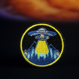 May include: A round, black embroidered patch with a yellow border. The patch features a blue UFO with a yellow beam of light shining down on a blue mountain range.