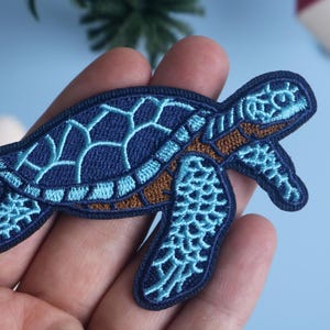 Può includere: Toppa ricamata a forma di tartaruga marina. La tartaruga ha un contorno e un guscio blu scuro con dettagli blu chiaro e marroni. La toppa è tenuta in mano su uno sfondo azzurro.
