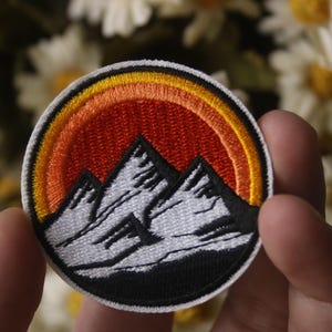 May include: Round embroidered patch featuring a mountain range under a sunset. The patch has a black border, white mountains, and a gradient of orange and yellow in the sky. The patch is held in a person's hand.