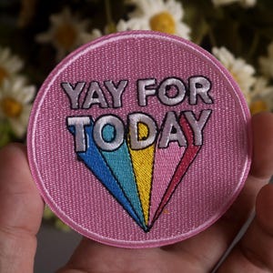 May include: A round, pink embroidered patch with the words "YAY FOR TODAY" in silver lettering. A colorful, triangular design in blue, yellow, and red is below the text. The patch is held in a hand.