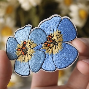 Puede incluir: Dos parches bordados de flores azules con centros amarillos y detalles naranjas. Los parches se sostienen en una mano.