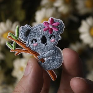 Pode incluir: Etiqueta bordada para passar a ferro com um coala cinzento com uma flor rosa na cabeça, agarrado a um ramo com folhas verdes.
