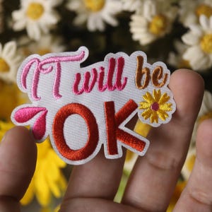Op de afbeelding: Geborduurde patch met de zin "It will be OK" in roze, oranje en gouden letters. De patch heeft een kleine gele bloem en wordt in een hand gehouden tegen een achtergrond van madeliefjes.