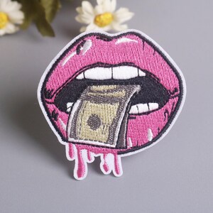Pode incluir: Patch bordado com uma boca com lábios rosa brilhantes e dentes brancos. Um maço de dinheiro está na boca, com gotas rosa por baixo. O patch tem uma borda branca e está em um fundo cinza.