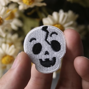 Op de afbeelding: Witte geborduurde strijk-op patch van een glimlachende schedel met een scheur in de bovenkant van zijn hoofd.