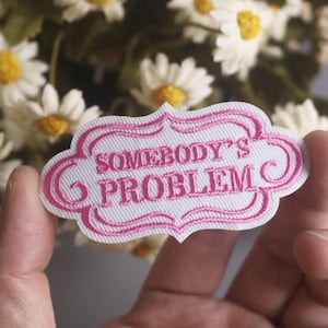 Può includere: Toppa bianca ricamata con bordi rosa e testo «SOMEBODY'S PROBLEM». La toppa ha un bordo decorativo. Lo sfondo include un bouquet di margherite bianche con centri gialli.
